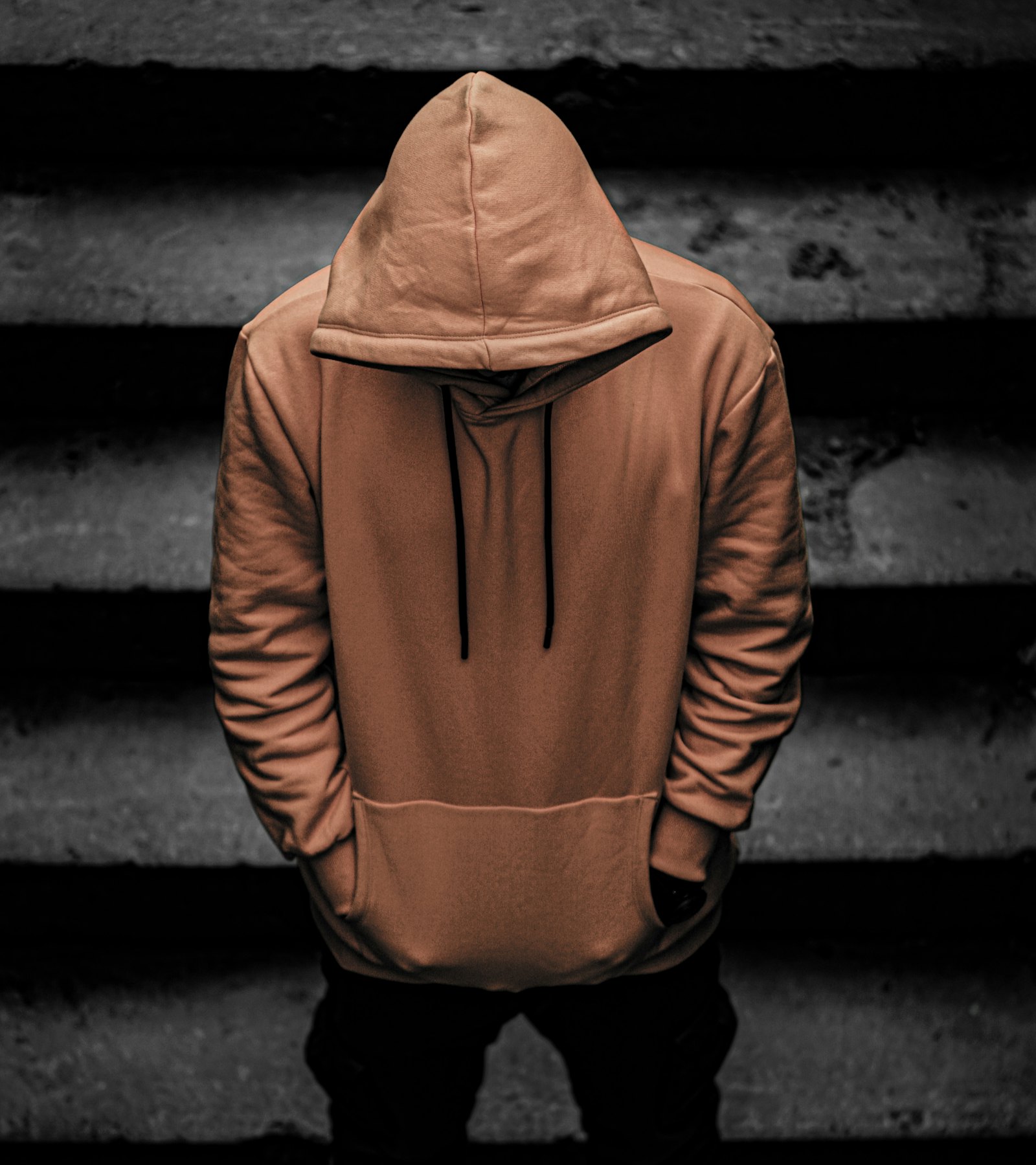 Persona de espaldas con sudadera de capucha color barro, cabeza inclinada, sobre unas escaleras de hormigón